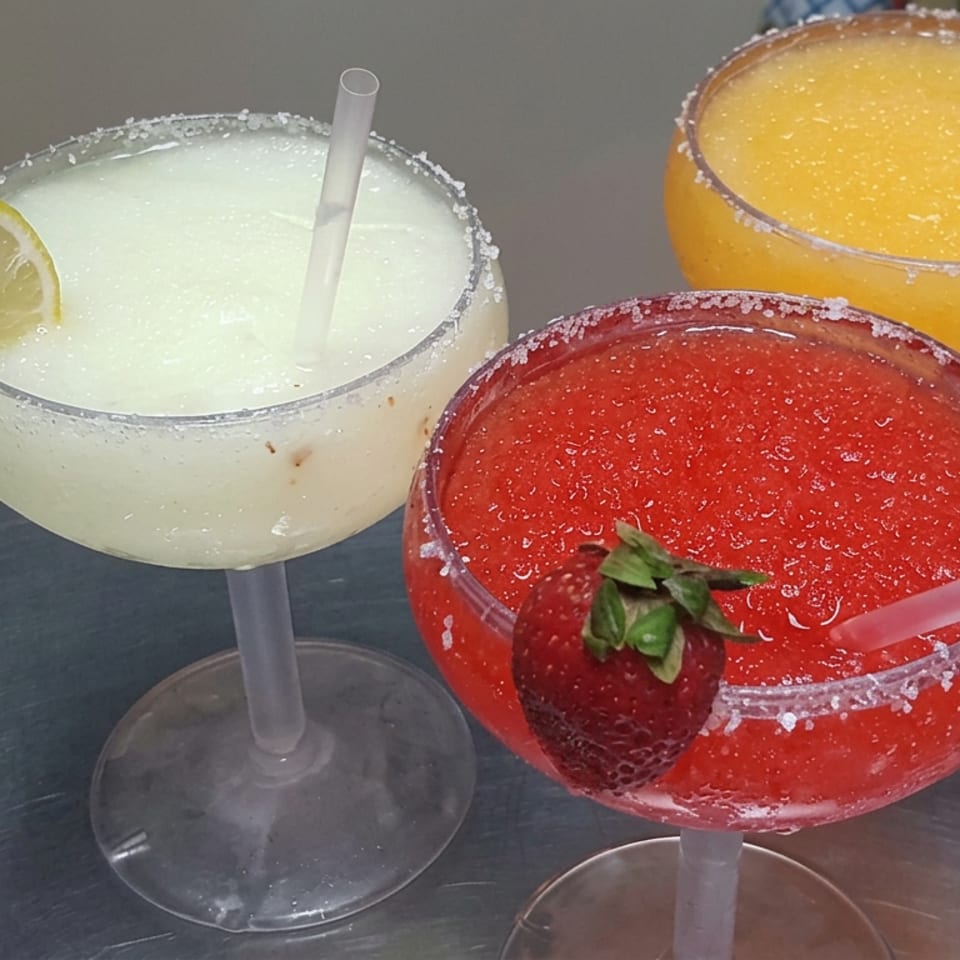 Three colorful frozen cocktails with salted rims, straws, and lemon and strawberry garnishes on a restaurant table.