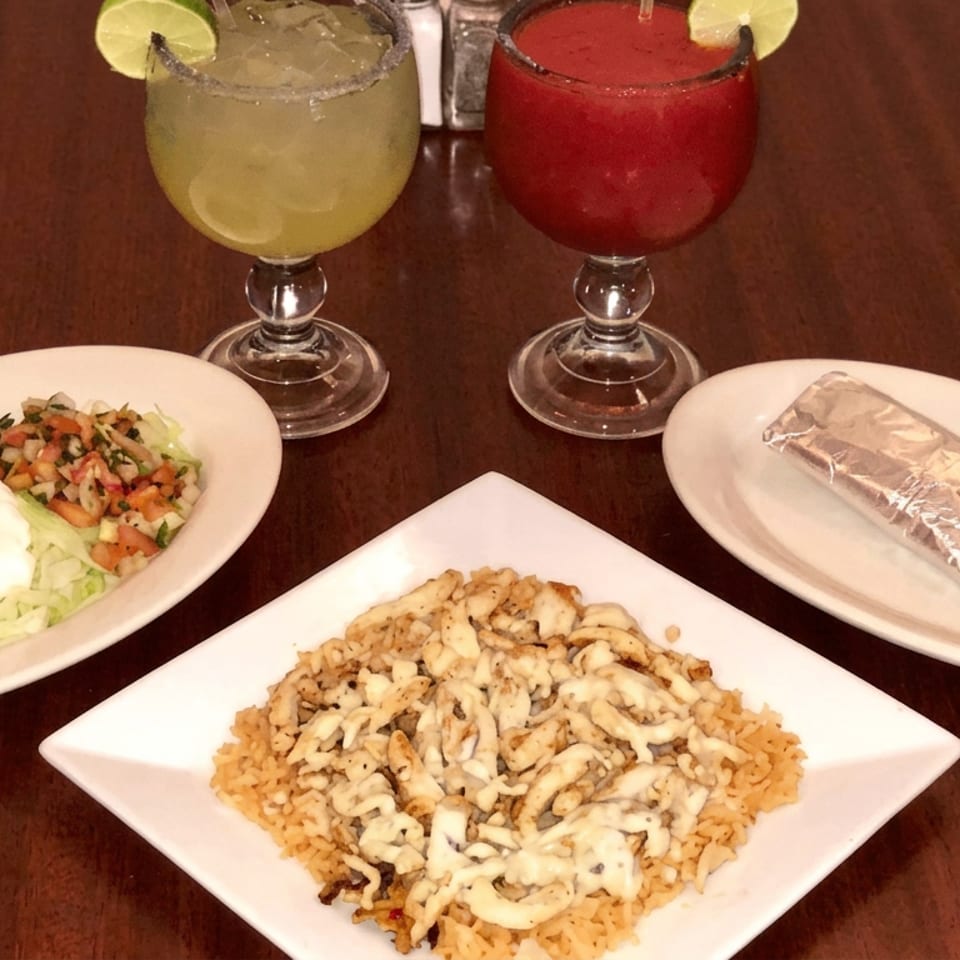 Restaurant table displays grilled chicken over rice, side salad, wrapped item, and four lime-garnished cocktails.