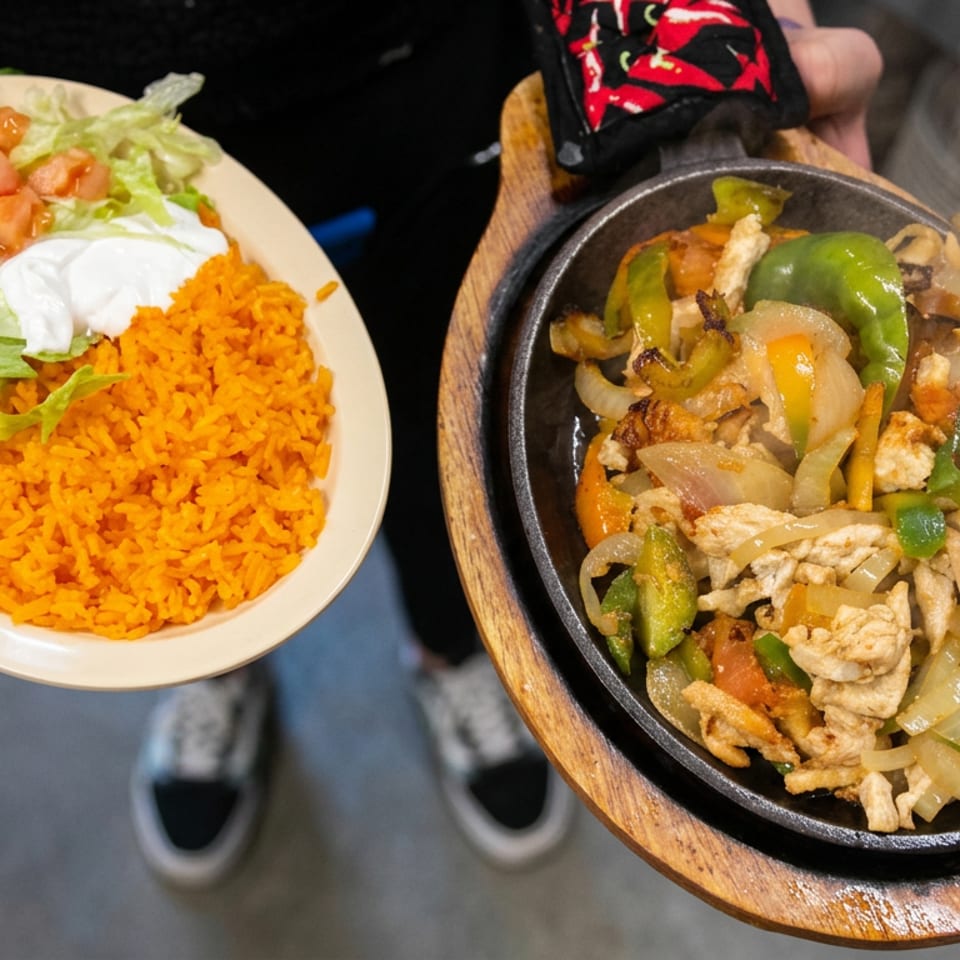 Fajita-style chicken with sautéed peppers and onions on a sizzling platter, served with Mexican rice, lettuce, tomatoes, and sour cream.
